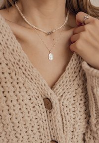 Gelaagde kettingen met een zilveren hanger met een boomontwerp, een parel choker en een textuurrijke, beige gebreide cardigan met houten knopen.