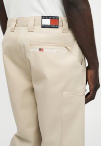 Μπεζ παντελόνι με τσέπες με φερμουάρ, κεντημένο λογότυπο της ομάδας USA SailGP κοντά στην πίσω τσέπη, και ετικέτα με το λογότυπο Tommy Hilfiger στη ζώνη της μέσης.