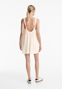 Helles beige ärmelloses Kleid mit lockerer Passform und Rückenschlitz, aus strukturiertem Stoff gefertigt. Kombiniert mit weißen Schuhen mit dunklen Akzenten.