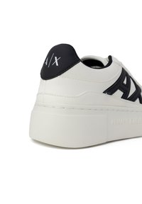 Sneaker bianca in pelle con accenti neri, caratterizzata da una suola platform spessa, logo rialzato sul tallone e texture liscia con elementi di design a contrasto.