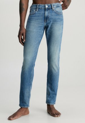 Barfodet person iført lyseblå falmede slim-fit jeans, der vises fra taljen til fødderne mod en ensfarvet lys baggrund.