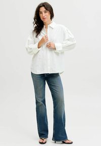Hvid button-up skjorte med krave, blussede ærmer og subtile prikmønstre; parret med flagrende blå denim jeans og sorte flip-flops.