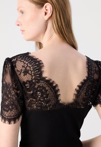 Haut noir avec des manches courtes en dentelle et un dos en V profond. La dentelle présente des motifs floraux et des bords frangés. Le corps est en tissu côtelé.