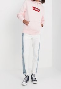 Sudadera rosa con el logo rojo de Levi's, combinada con unos jeans blancos que tienen paneles laterales en azul claro. Unos zapatillas negras altas completan el look.