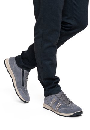 Person wearing dark pants and gray lace-up sneakers with side stripes and zipper detail, standing with one foot crossed over the other.