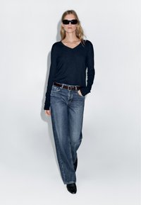 Navy long-sleeve V-neck top paired with loose-fitting, faded blue jeans and a brown belt. Model wearing dark sunglasses and black boots.