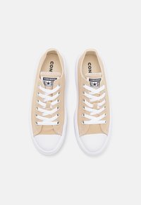 Beiga canvas sneakers med vita gummisulor, med vita snörningar, metallöglor och en svart "Converse All Star"-logotyp på tungan.