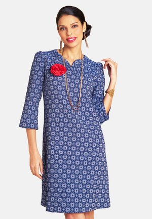 Woman wearing a blue patterned dress with three-quarter sleeves, a red flower brooch, gold long necklace, and matching earrings and bracelet.