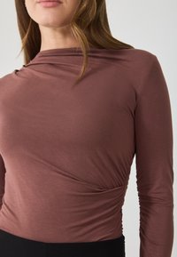 Garais piedurkņu tops bālganā mauve krāsā, ar krītošu dekoltē un savilktu sānu detaļu, izgatavots no mīksta, stiepjama auduma.