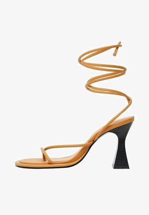 Sandalia de tacón alto de color naranja con tiras, lazo en el tobillo en espiral, suela plana y un tacón en bloque negro de forma geométrica. Textura suave en toda la sandalia.