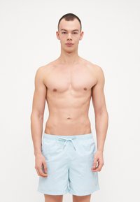 Lys blå badeshorts laget av lett stoff, med elastisk midje og snor, vist på en person uten topp.