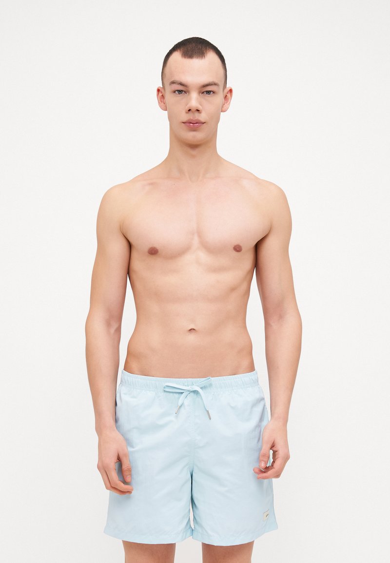 Lys blå badeshorts laget av lett stoff, med elastisk midje og snor, vist på en person uten topp.