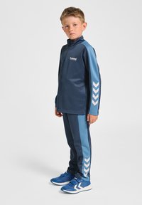 Marineblauer sportlicher Trainingsanzug mit einem Halb-Reißverschluss-Oberteil, blauen Seitenpanelen und weißen Chevron-Details, kombiniert mit passenden Hosen und blauen Schuhen.