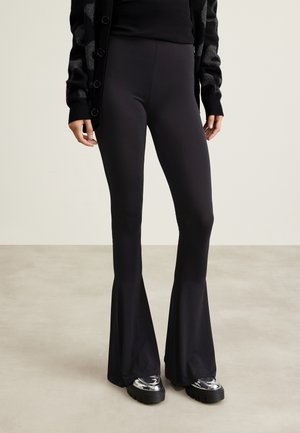 Persona con pantalones negros ajustados acampanados, cárdigan negro con estampado y zapatos metálicos de plataforma gruesa sobre un suelo claro y fondo de pared lisa.