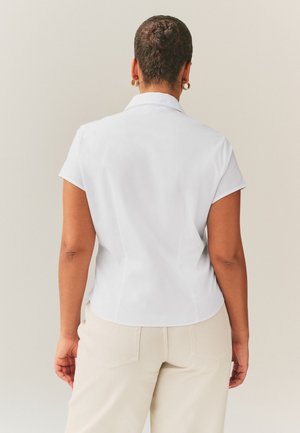 Žena s copánkem ve tvaru drdolu, nosí zlaté kruhové náušnice, bílou košili s krátkým rukávem a límečkem a béžové kalhoty, otočená zády k jednoduchému pozadí.