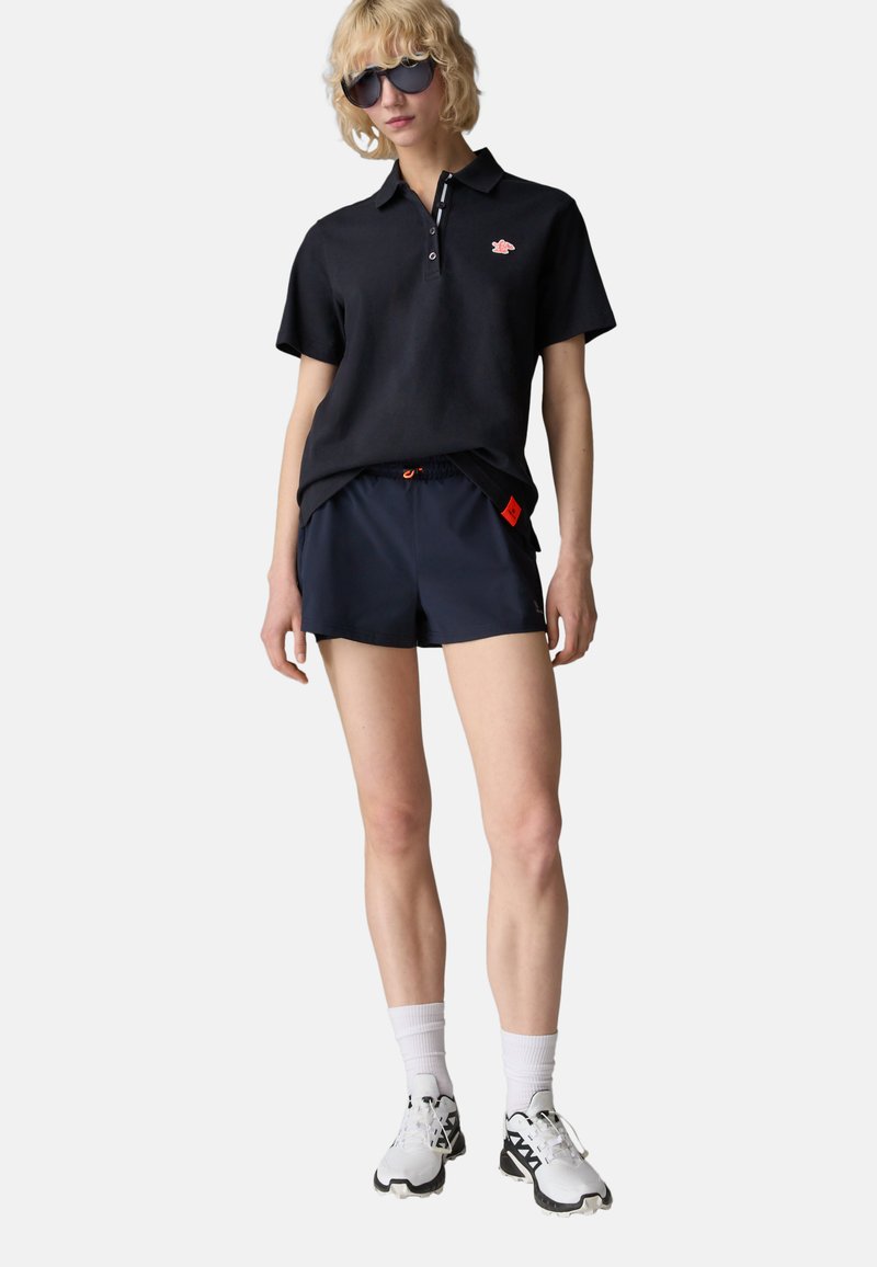 Schwarzes Poloshirt mit einem Logo, marineblaue Shorts, weiße Sneakers und Sonnenbrille. Das Outfit besteht aus atmungsaktiven Materialien und hat einen lockeren Schnitt.