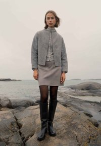 Giacca bomber di lana grigia, top grigio chiaro con bottoni, gonna grigia, stivali neri fino al ginocchio e collant neri trasparenti su una costa rocciosa.