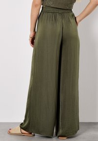 Une personne se tient de dos, portant un pantalon large vert olive et un haut assorti, avec des sandales brunes aux pieds. Fond et sol clairs.