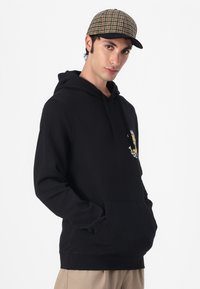 Felpa nera con tasca a marsupio, cappuccio con coulisse e una grafica colorata sul petto. Berretto piatto con motivo a quadretti in beige e marrone.