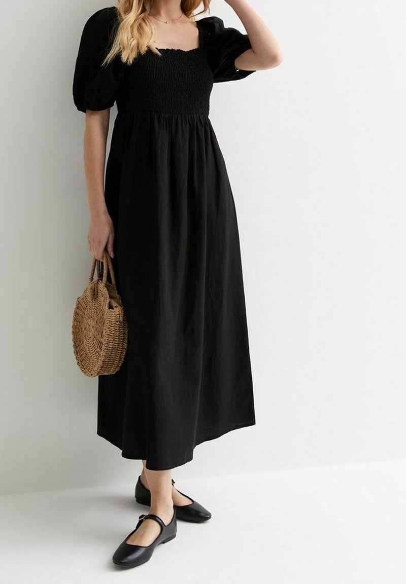 Femme portant une robe midi noire à manches bouffantes, tenant un sac rond en paille tressée et portant des chaussures Mary Jane noires.