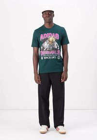 Green cotton t-shirt featuring large "ADIDAS ORIGINALS" print in pink and gold, paired with black pants and white sneakers with red stripes.
