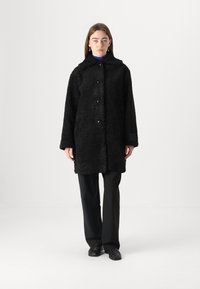 Cappotto nero in pelliccia morbida con colletto e chiusura a bottone. Presenta maniche lunghe e una vestibilità rilassata, indossato sopra un top a strati viola.