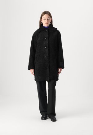 Cappotto nero in pelliccia morbida con colletto e chiusura a bottone. Presenta maniche lunghe e una vestibilità rilassata, indossato sopra un top a strati viola.