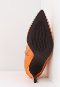 Chaussure pointue en cuir orange avec un talon noir fin, reposant semelle vers le haut contre un coin blanc en arrière-plan.