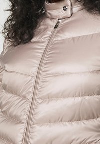 Beige pufferjacka med en glänsande, slät textur. Har en hög krage och en framzipper, med kviltade sektioner för isolering.