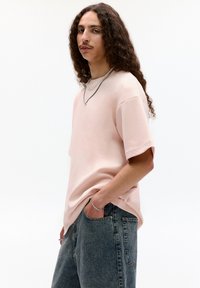 Világos rózsaszín, oversized póló rövid ujjakkal és kerek nyakkal, elmosódott kék farmernadrággal párosítva. Kiegészítők között rétegelt ezüstláncok találhatók.