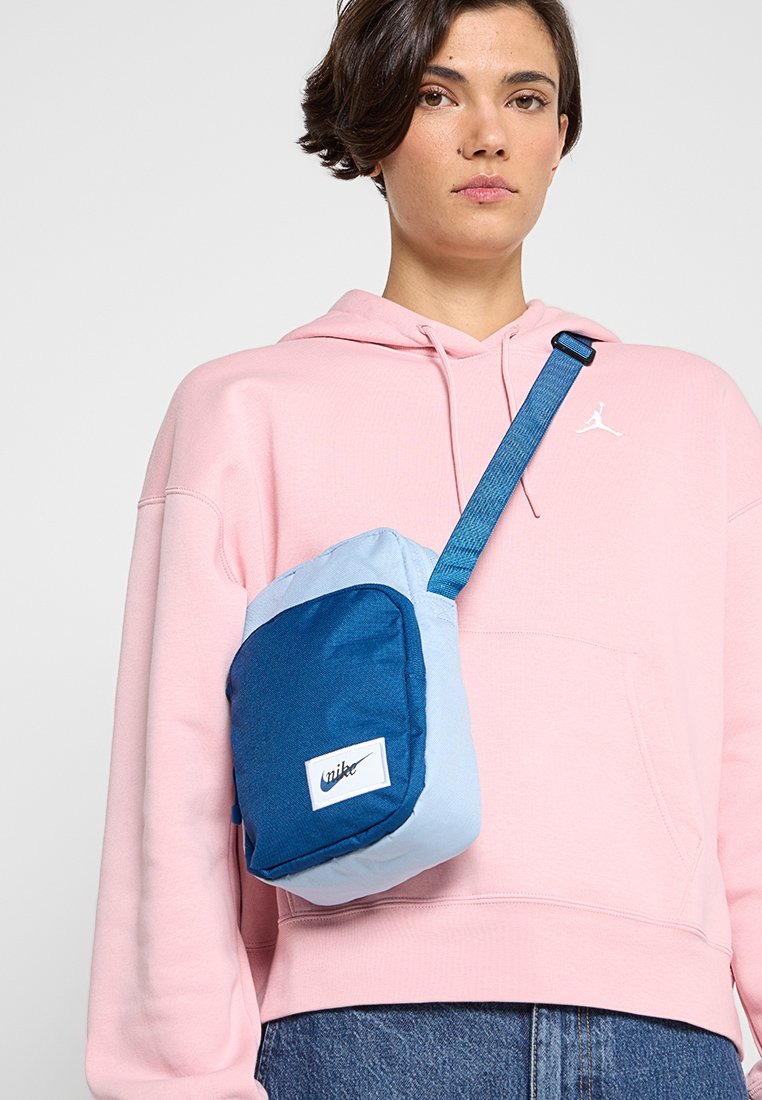 Bolso pequeño para el hombro en materiales azul y azul claro; cuenta con un parche de logo blanco y una correa ajustable. Llevado sobre una sudadera con capucha rosa.