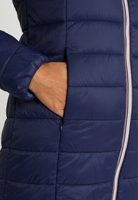 Chaqueta acolchada azul marino con textura acolchada, cremallera de longitud completa y bolsillo lateral. Presenta un ribete rosa contrastante a lo largo de la cremallera.