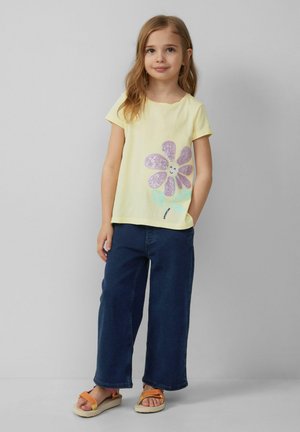 Geel T-shirt met korte mouwen en paars glinsterende bloemenprint. Gecombineerd met donkerblauwe wijde broek en oranje sandalen. Glad textuur van de stof.