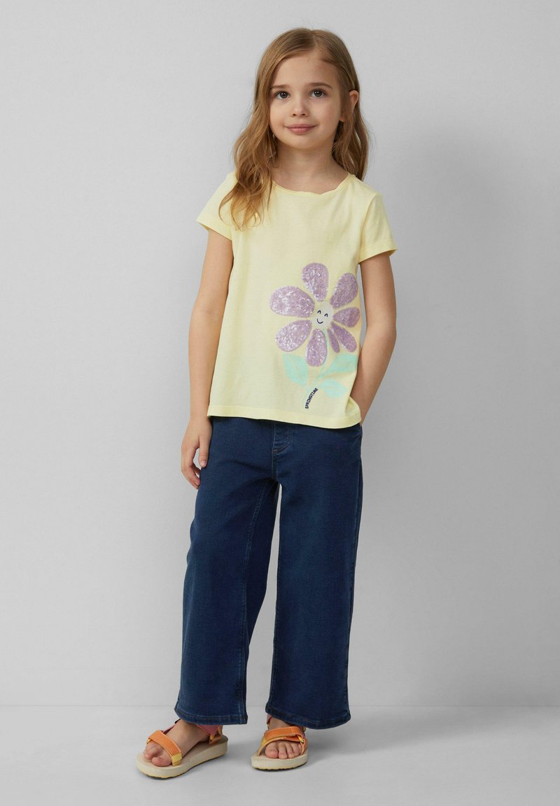 Geel T-shirt met korte mouwen en paars glinsterende bloemenprint. Gecombineerd met donkerblauwe wijde broek en oranje sandalen. Glad textuur van de stof.