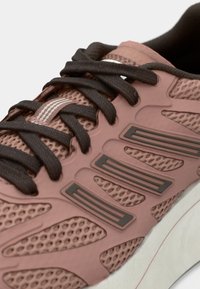 Lyserosa idrettssko med pustende mesh-overdel, mørkebrune snører og teksturerte gummidetaljer på sidene. Buet hvit såle.