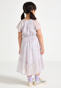 Robe violet clair avec des motifs floraux, jupe à plusieurs couches, taille élastique et manches courtes évasées. Associée à des baskets argentées et des chaussettes violettes.