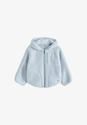 Lächelndes Mädchen mit langem blondem Haar, das eine hellblaue, flauschige Jacke mit Kapuze und blaue Cargohosen trägt und mit den Händen in den Taschen steht.