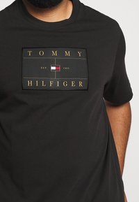 Camiseta de algodón negra con un gráfico rectangular que presenta "TOMMY HILFIGER" en dorado y un pequeño detalle de bandera en rojo, blanco y azul.