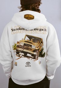 Fehér kapucnis pulóver, amelyen egy vintage Toyota Land Cruiser grafika látható, a hátoldalán "LAND CRUISER" felirattal és egy logóval, kiemelve egy barna jármű illusztrációját.
