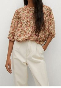 Eine Frau trägt eine beigefarbene Bluse mit floralen Mustern und Puffärmeln, die in hochgeschnittene cremefarbene Hosen gesteckt ist. Eine Hand steckt in der Tasche und ihr langes, dunkles Haar ist sichtbar.