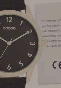 Montre ronde avec un cadran noir, des chiffres arabes blancs et un boîtier beige. Équipée d'un bracelet noir et inclut une étiquette de conformité.