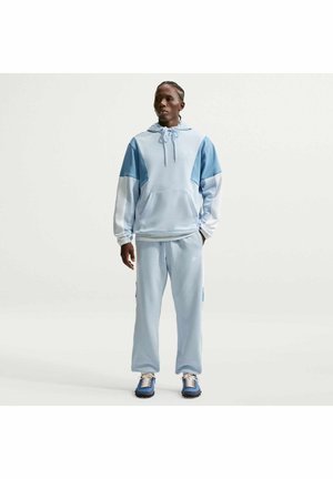 Uomo in piedi rivolto in avanti con felpa con cappuccio a blocchi di colore azzurro e bianco, pantaloni della tuta coordinati e scarpe da ginnastica blu, su sfondo semplice.