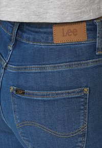 Jeans de mezclilla azul con un parche de cuero marrón etiquetado "Lee" en la cintura. Presenta costuras amarillas y un diseño estándar de bolsillo trasero.
