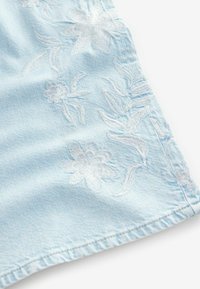 Lichtblauwe denimstof met witte bloemenborduring en bladvormen. Heeft een gestikte zoom en een zachte textuur.
