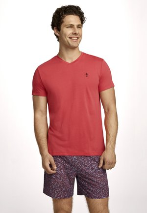 Smiling man wearing a red V-neck T-shirt and patterned shorts, standing against a plain light background.