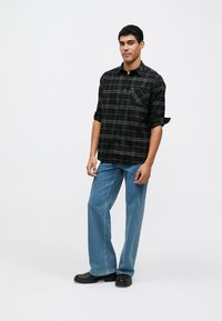 Black and gray plaid shirt with a single chest pocket, paired with blue straight-leg jeans and black boots, standing against a white background.