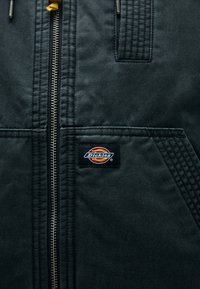 Μαύρο ζακέτα με φερμουάρ, κατασκευασμένη από υφασμάτινη υφή, με εμφανή ραφή και εμφανές λογότυπο "Dickies" κοντά στο φερμουάρ.