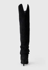 Botas de ante negras hasta la rodilla con un corte en forma de V en la parte superior, tacón alto en forma de cono y diseño suelto para añadir textura.
