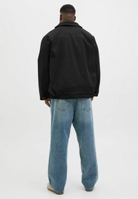 Sort jakke med krave, afslappet pasform og elastisk bund, kombineret med lyseblå viddejeans. Baggrunden er helt hvid.