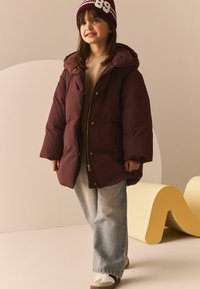 Jachetă puffer bordeaux cu glugă, fermoar auriu și nasturi cu capse; asociată cu blugi de culoare bleu deschis și adidași albi, cu accente întunecate.